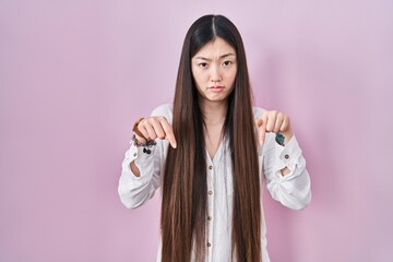 Chinese young woman standing over pink background pointing down looking sad and upset, indicating direction with fingers, unhappy and depressed.