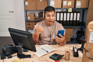Hispanic young man working at small business ecommerce on video call pointing thumb up to the side smiling happy with open mouth