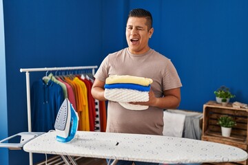 Hispanic young man holding folded laundry after ironing winking looking at the camera with sexy...
