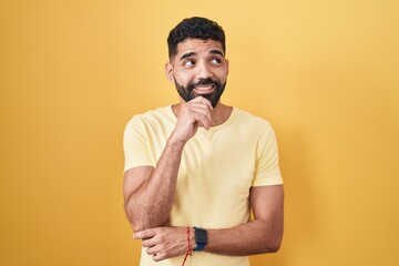 Hispanic man with beard standing over yellow background with hand on chin thinking about question, pensive expression. smiling and thoughtful face. doubt concept.