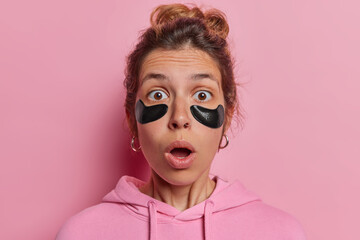 Beauty model attaches black hydrogel patches undergoes daily skincare routine has widely opened mouth reacts to something shocking dressed in sweatshirt isolated over pink studio background.