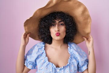 Fototapeta premium Young brunette woman with curly hair wearing summer hat looking at the camera blowing a kiss being lovely and sexy. love expression.
