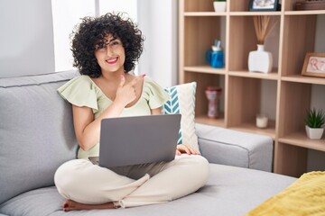 Young brunette woman with curly hair using laptop sitting on the sofa at home cheerful with a smile of face pointing with hand and finger up to the side with happy and natural expression on face
