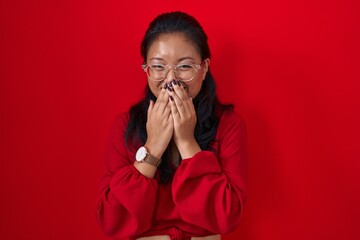 Asian young woman standing over red background laughing and embarrassed giggle covering mouth with hands, gossip and scandal concept