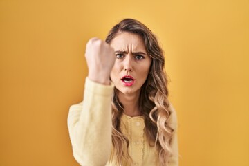 Young caucasian woman standing over yellow background angry and mad raising fist frustrated and furious while shouting with anger. rage and aggressive concept.