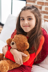 Adorable hispanic girl hugging teddy bear sitting on bed at bedroom