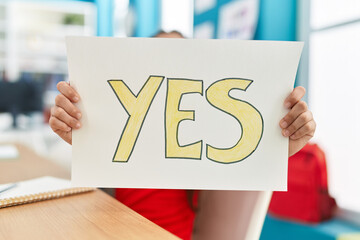 Adorable hispanic girl student sitting on table holding yes paper at classroom