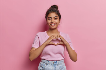 Portrait of happy Indian woman with hair bun shows finger heart gesture sends love to someone smiles pleasantly wears casual clothes isolated on pink background expresses sympathy feels passionate - 604670918