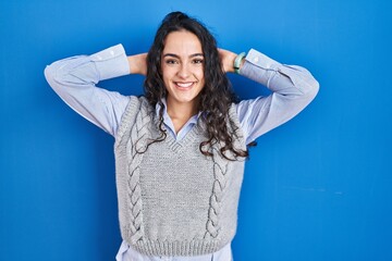 Young brunette woman standing over blue background relaxing and stretching, arms and hands behind head and neck smiling happy