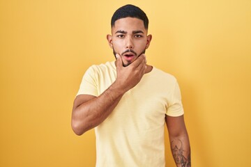 Young hispanic man standing over yellow background looking fascinated with disbelief, surprise and amazed expression with hands on chin
