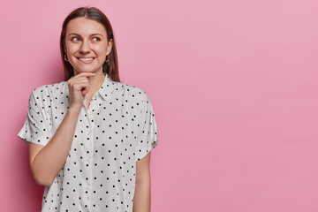 Fototapeta na wymiar Good looking cheerful female model with dark straight hair bites lips keeps hand on chin wears polka dot blouse has dreamy expression isolated over pink background copy space for your advertisement