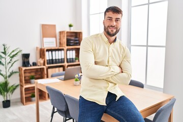 Young hispanic man business worker sitting with arms crossed gesture at office