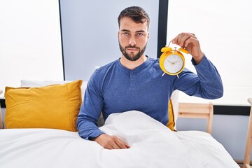 Handsome hispanic man in the bed holding alarm clock thinking attitude and sober expression looking self confident