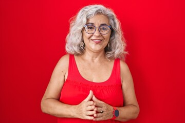 Middle age woman with grey hair standing over red background hands together and fingers crossed smiling relaxed and cheerful. success and optimistic