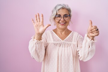 Middle age woman with grey hair standing over pink background showing and pointing up with fingers number six while smiling confident and happy.