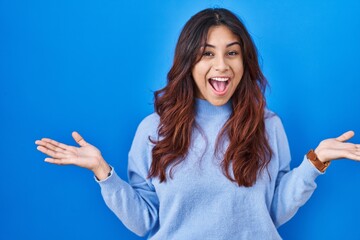 Fototapeta premium Hispanic young woman standing over blue background celebrating crazy and amazed for success with arms raised and open eyes screaming excited. winner concept