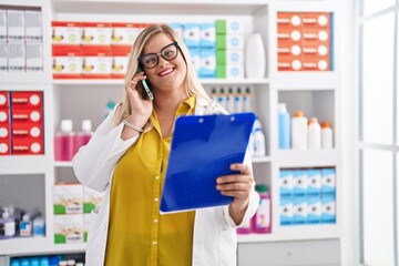 Young woman pharmacist talking on smartphone reading document at pharmacy