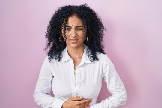 Hispanic woman with curly hair standing over pink background with hand on stomach because indigestion, painful illness feeling unwell. ache concept.