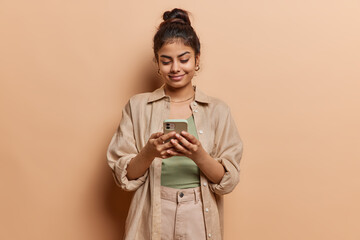 Indoor shot of beautiful charming young Indian woman uses smartphone for chatting online types message dressed in casual shirt and trousers browses social media isolated over brown background.