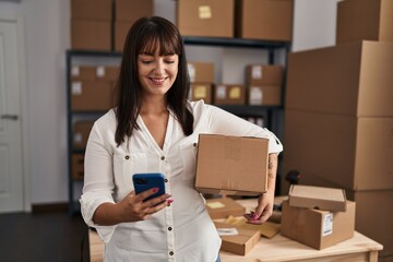 Fototapeta na wymiar Young beautiful hispanic woman ecommerce business worker using smartphone holding package at office