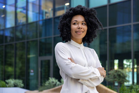 Confident adult mature businesswoman outside urban office business building, African American boss with crossed arms looking away, female worker in shirt satisfied with achievement results at work.