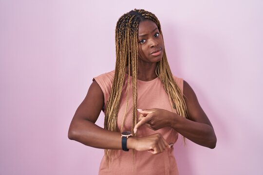 African american woman with braided hair standing over pink background in hurry pointing to watch time, impatience, upset and angry for deadline delay