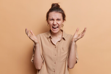 Angry fed up young European woman spreads palms and yells annoyed argues with someone wears shirt expresses negative emotions isolated over brown background. How dare you. Furious female model