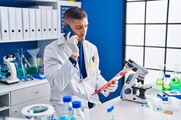 Young hispanic man scientist talking on the smartphone reading document at laboratory