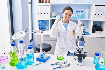 Fototapeta premium Young brunette woman working at scientist laboratory very happy and excited doing winner gesture with arms raised, smiling and screaming for success. celebration concept.