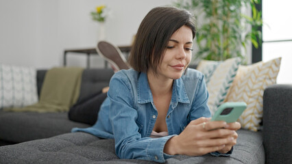 Obraz premium Young caucasian woman using smartphone lying on sofa at home