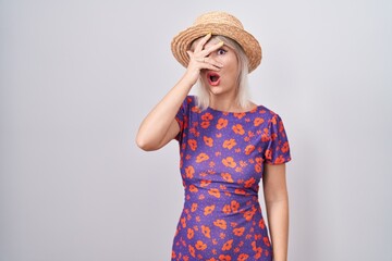 Young caucasian woman wearing flowers dress and summer hat peeking in shock covering face and eyes with hand, looking through fingers with embarrassed expression.