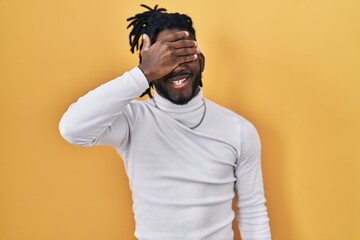 African man with dreadlocks wearing turtleneck sweater over yellow background smiling and laughing with hand on face covering eyes for surprise. blind concept.