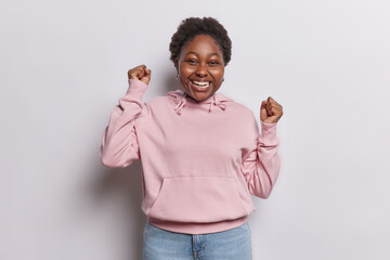 Portrait of excited dark skinned woman celebrates lucky win cheers and raises clenched fists up...