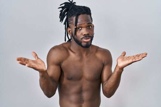 African man with dreadlocks standing shirtless over isolated background clueless and confused expression with arms and hands raised. doubt concept.