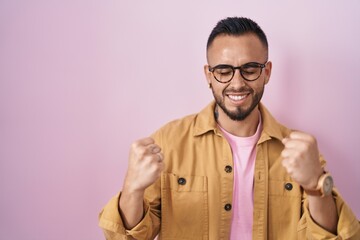 Fototapeta premium Young hispanic man standing over pink background very happy and excited doing winner gesture with arms raised, smiling and screaming for success. celebration concept.