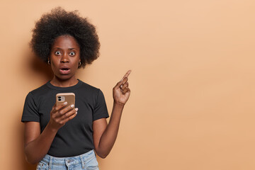 Horizontal shot of impressed black woman with bushy hair holds mobile phone reacts to something astonishing holds mobile phone dressed in black t shirt and jeans isolated over brown background - obrazy, fototapety, plakaty