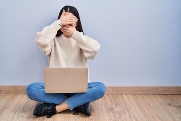 Young woman using laptop sitting on the floor at home covering eyes and mouth with hands, surprised and shocked. hiding emotion