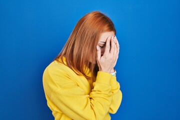 Young woman standing over blue background with sad expression covering face with hands while...