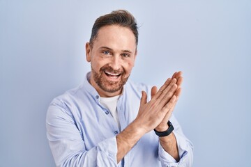 Middle age caucasian man standing over blue background clapping and applauding happy and joyful, smiling proud hands together