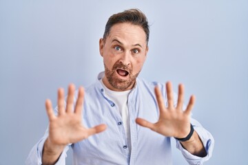 Fototapeta premium Middle age caucasian man standing over blue background afraid and terrified with fear expression stop gesture with hands, shouting in shock. panic concept.