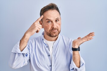 Middle age caucasian man standing over blue background confused and annoyed with open palm showing copy space and pointing finger to forehead. think about it.
