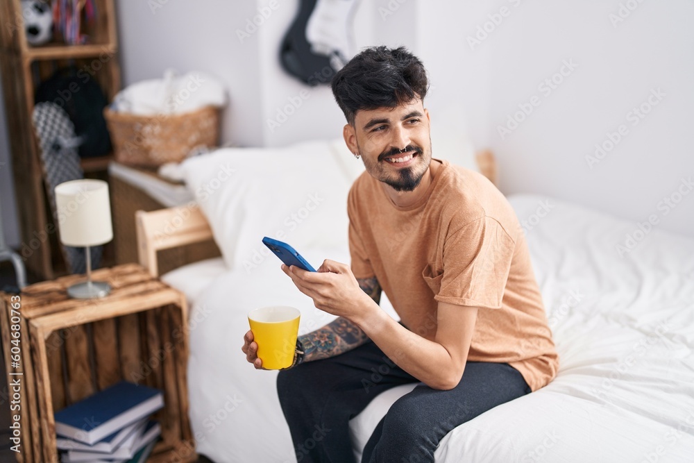 Wall mural Young hispanic man using smartphone drinking coffee at bedroom