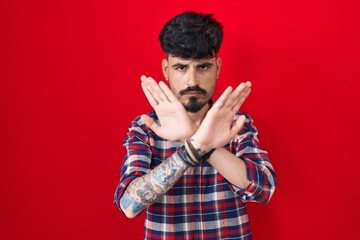 Young hispanic man with beard standing over red background rejection expression crossing arms and palms doing negative sign, angry face