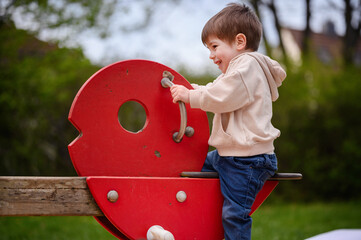 Little toddler boy in beige stylisch hoodie and jeans swinging on red seesaw on playground