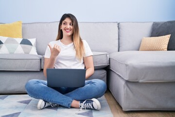 Young brunette woman sitting on the floor at home using laptop pointing thumb up to the side...