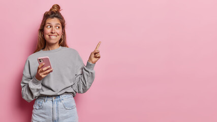 Studio shot of pretty millennial girl bites lips points index finger aside on blank space uses smartphone recommends you to click on link dressed in fashionable clothes isolated over pink background