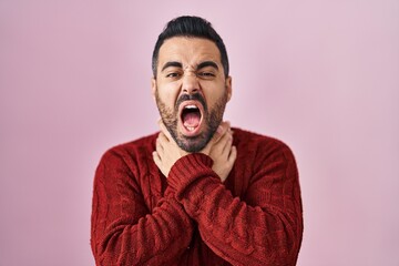 Young hispanic man with beard wearing casual sweater over pink background shouting and suffocate because painful strangle. health problem. asphyxiate and suicide concept.