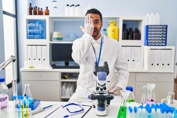 Fototapeta premium Young hispanic man with beard working at scientist laboratory covering one eye with hand, confident smile on face and surprise emotion.