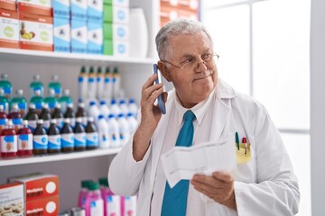 Middle age grey-haired man pharmacist talking on smartphone reading prescription at pharmacy