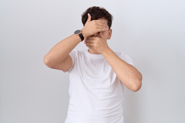 Young non binary man wearing casual white t shirt covering eyes and mouth with hands, surprised and shocked. hiding emotion